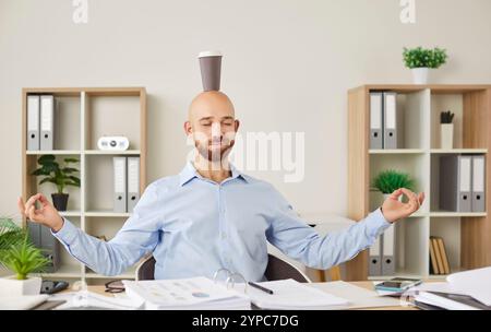 Ruhiger, lustiger, ruhiger Büroangestellter, der an der Rezeption sitzt und entspannend Yoga Mudra mit einer Tasse auf dem Kopf macht Stockfoto