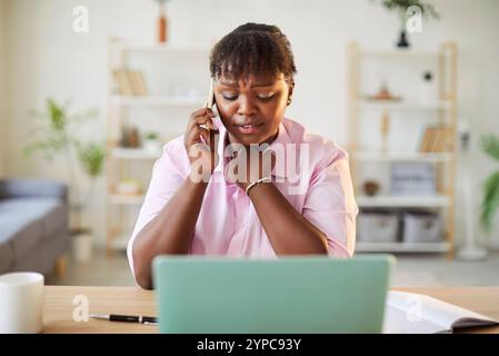 Verärgerte Geschäftsfrau, die an einem Laptop arbeitet und mit dem Mobiltelefon spricht, hat unangenehme Gespräche. Stockfoto