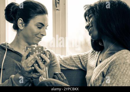 Zwei Frauen lächeln einander an und genießen einen gemütlichen Moment bei Kaffee. Schwarzweißfoto für eine warme und intime Verbindung Stockfoto