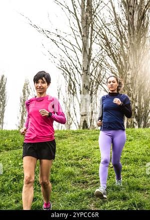 Zwei Frauen joggen auf einem grasbewachsenen Parkweg, umgeben von Bäumen. Konzentrieren Sie sich auf eine Outdoor-Fitness-Aktivität im Frühling, um Gesundheit, Bewegung und zu fördern Stockfoto