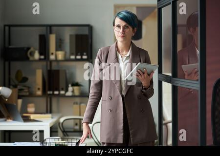 Selbstbewusste Geschäftsfrau mittleren Alters mit Tablet, die die Kamera ansieht, während sie Online-Finanzdaten im Büro analysiert Stockfoto