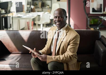 Selbstbewusster, reifer afroamerikanischer Geschäftsmann mit Finanzdokumenten in der Hand, der Sie während der Arbeit auf der Couch ansieht Stockfoto