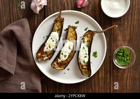 Gebackene Aubergine mit Knoblauch-Joghurtdressing auf weißem Teller auf hölzernem Hintergrund. Draufsicht, flach Stockfoto