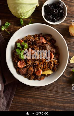 Gedünsteter Kohl (polnische Bigos) mit Sauerkraut, Pilzen, Räucherfleisch und Gewürzen in einer Schüssel auf Holztisch. Draufsicht, flach Stockfoto