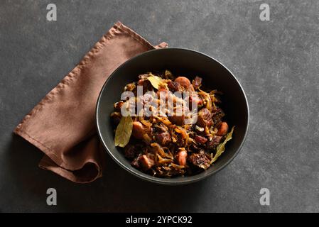 Polieren Sie Bigos in der Schüssel auf dunklem Steinhintergrund. Gedünsteter Kohl mit Sauerkraut, Pilzen, Räucherfleisch und Gewürzen. Nahansicht Stockfoto