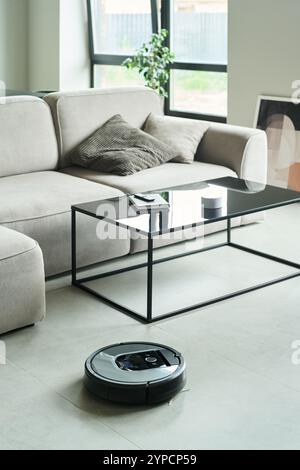 Roboter-Staubsauger, der in einem modernen Wohnzimmer mit Kaffeetisch aus Glas, beigefarbenem Sofa und großen Fenstern funktioniert. Gemütliches Ambiente mit Kissen und gerahmtem Kunstwerk, das im Hintergrund sichtbar ist Stockfoto