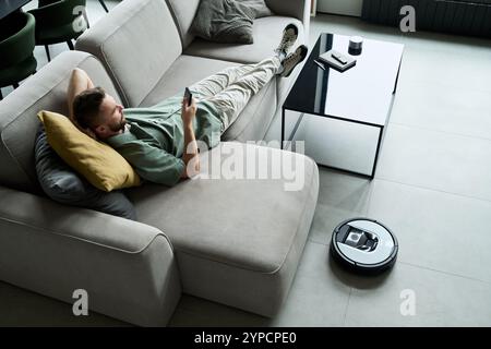 Mann, der auf dem Sofa liegt und Smartphone neben dem Kaffeetisch mit Roboter-Vakuum auf dem Boden in der Nähe surft und eine moderne und entspannte Atmosphäre im Wohnzimmer schafft Stockfoto