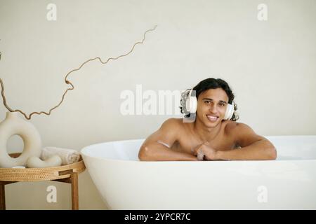Porträt eines lächelnden jungen erwachsenen Mannes mit Kopfhörern auf dem Kopf, der sich beim Posen auf das Bad lehnt Stockfoto