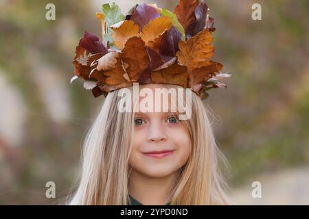 Entzückendes kleines Mädchen mit langen blonden Haaren und blauen Augen, das eine Krone aus trockenen Herbstblättern trägt. Kinder haben eine Idee mit Herbstlaub. Stockfoto