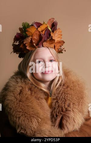 Entzückendes kleines Mädchen mit langen blonden Haaren und blauen Augen, das eine Krone aus trockenen Herbstblättern trägt. Kinder haben eine Idee mit Herbstlaub. Stockfoto