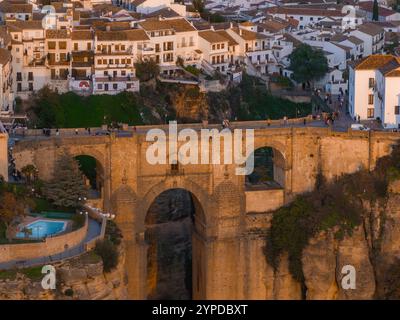 Das Bild zeigt eine Luftaufnahme von Puente Nuevo in Ronda, Spanien, mit weiß getünchten Gebäuden auf Klippen. Der Sonnenuntergang wirft ein goldenes Leuchten über die Szene. Stockfoto