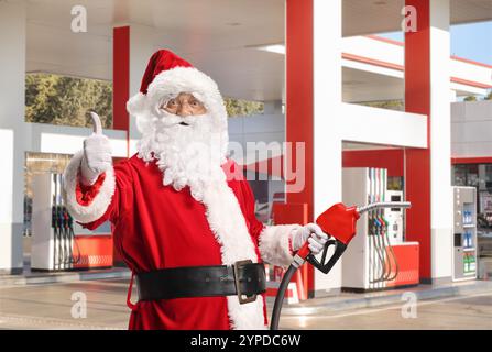 Der Weihnachtsmann hält eine Kraftstoffeinspritzdüse und drückt die Daumen an einer Tankstelle hoch Stockfoto