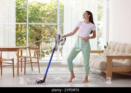 Lächelnde junge Frau, die zu Hause mit schnurlosem Staubsauger den Boden reinigt Stockfoto