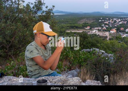 Kleiner Junge mit Brille und Mütze, der an Sommertagen Fotos in der Natur mit Blick auf das malerische Dorf macht. Konzept der kindlichen Erkundung, Fotografie h Stockfoto