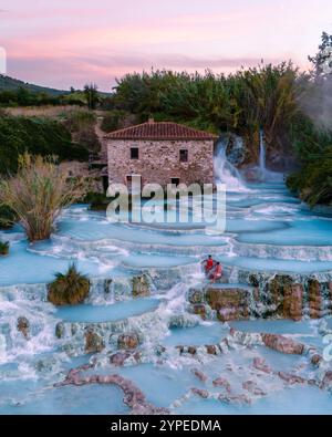 Besucher genießen das warme, mineralreiche Wasser der Thermalbäder von Saturnia, umgeben von üppigem Grün und kaskadierenden Wasserfällen, während die Sonne untergeht und den Himmel in atemberaubenden Rosa- und Orangetönen malt. Stockfoto