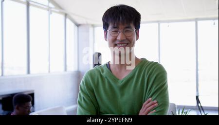 Vorderansicht eines jungen gutaussehenden asiatischen Managers, der mit überkreuzten Armen im modernen Büro steht Stockfoto