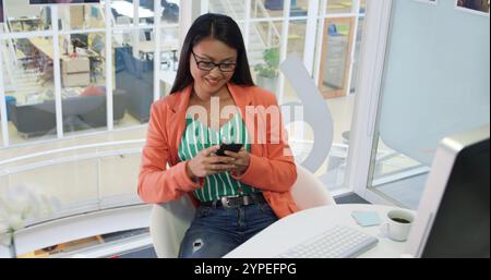 Vorderansicht einer jungen, birassischen Geschäftsfrau, die in einem modernen Büro arbeitet, mit einem Smartpho Stockfoto