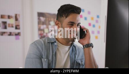 Vorderansicht eines glücklichen, birassischen Mannes, der in einem kreativen Büro arbeitet, auf dem Smartphone spricht und schaut Stockfoto