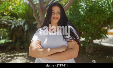 Junge Frau mit langen Haaren, die in einem Stadtpark steht, mit überkreuzten Armen, selbstbewusst im Schatten der Bäume lächelt. Stockfoto