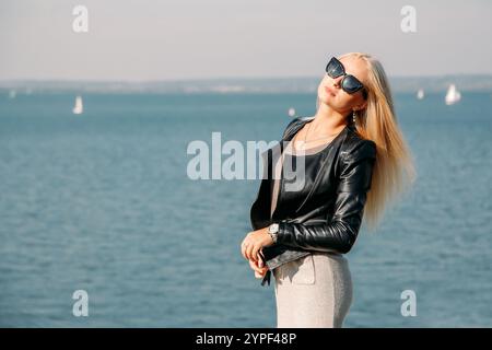 Junge Frau mit langen blonden Haaren, Sonnenbrille und Lederjacke, posiert am Meer Stockfoto