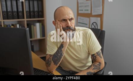 Hispanischer Mann mit Bart und Tätowierungen sitzt drinnen am Schreibtisch, tief in Gedanken, umgeben von Regalen mit Ordnern und Pinnwänden. Stockfoto