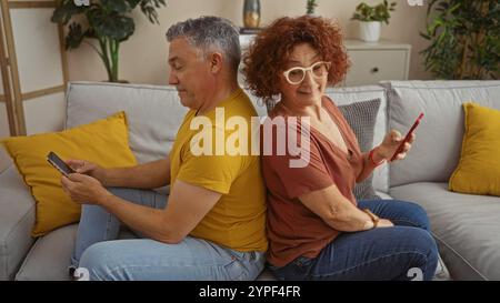 Ein Paar mittleren Alters sitzt hinten auf einem Sofa zu Hause, benutzt Smartphones, während die Frau in einem gemütlichen li hinterhältig auf den Bildschirm des Mannes blickt Stockfoto