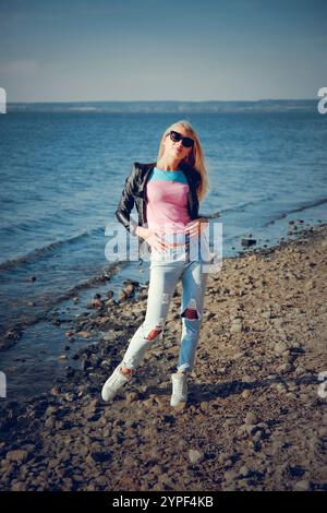 Blondes Model mit Sonnenbrille, zerrissener Jeans, rosa T-Shirt und schwarzer Lederjacke posiert an einem felsigen Strand Stockfoto