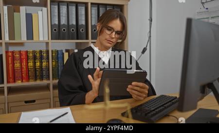 Eine junge Richterin in einem Gerichtssaal, die Dokumente auf einem Tablet überprüft, umgeben von Rechtsbüchern und Büromaterialien, die eine professionelle umwelt anzeigen Stockfoto