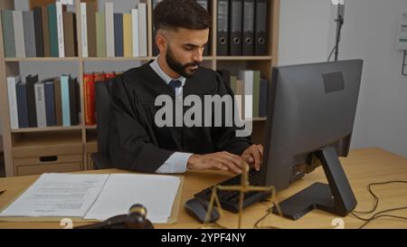 Junger hispanischer Mann mit Bart, der in einem Büro als Richter arbeitet, einen schwarzen Mantel trägt und an einem Computer mit juristischen Dokumenten und einem Hammer auf dem Th schreibt Stockfoto