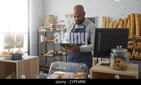 Junger Mann im Bäckereiraum drinnen mit Glatzkopf und Bart, der Tablette benutzt, während er hinter der Theke in blauer Schürze steht Stockfoto