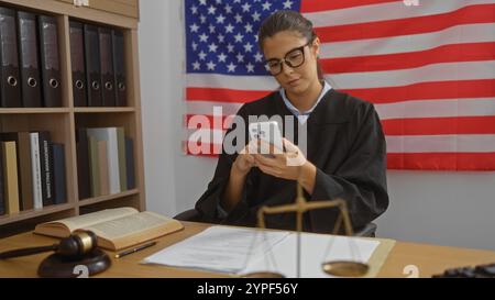 Richterin mit brünetten Haaren, die in einem Gerichtssaal an einem Smartphone arbeitet, mit amerikanischer Flagge, Gesetzbüchern und Richtergabel auf dem Schreibtisch Stockfoto