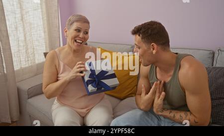 Die Mutter empfängt glücklich ein Geschenk von ihrem Sohn im Wohnzimmer mit einem großen Lächeln, zeigt ihre starke Familienbindung und Liebe, sitzt zusammen auf einem Sof Stockfoto