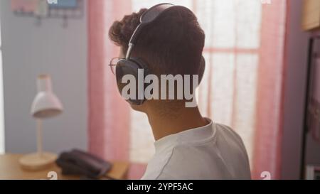 Junger hispanischer Mann in einem Büro mit Kopfhörern, von hinten gesehen, mit Lampe und Telefon auf dem Schreibtisch in einem hell beleuchteten Innenarbeitsbereich. Stockfoto