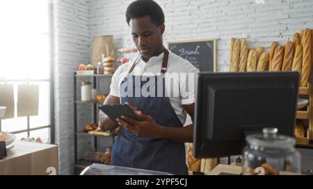 Junger Mann, der in einer Bäckerei mit einer Tablette arbeitet, umgeben von Gebäck, Brotregalen und einer modernen Kasse Stockfoto
