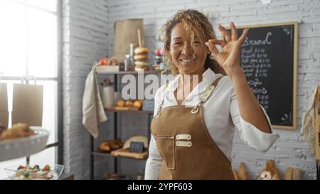Frau lächelt in der Bäckerei und zeigt eine gute Geste mit Gebäck auf Regalen im Hintergrund Stockfoto