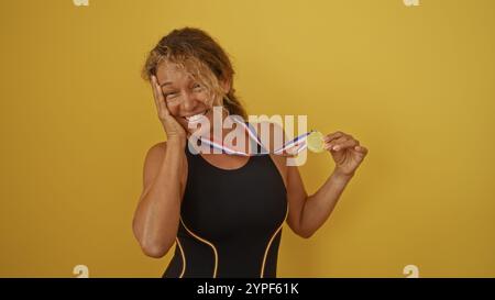 Die hispanische Frau hält stolz eine Goldmedaille mit fröhlichem Ausdruck, die vor einem leuchtend gelben Hintergrund steht. Stockfoto