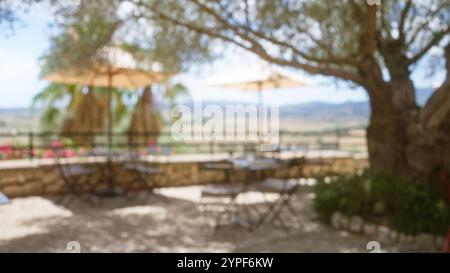 Unscharfe Outdoor-Szene auf mallorca mit unscharfen Tischen, Stühlen und Sonnenschirmen unter Bäumen mit Blick auf die ferne Landschaft vor blauem Himmel und Stockfoto