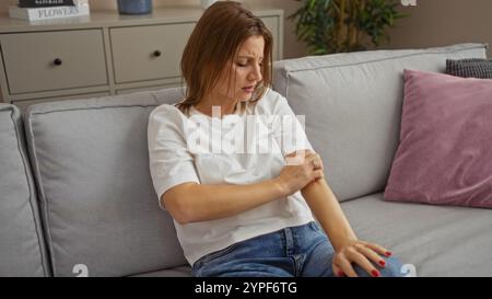 Eine junge, attraktive Frau sitzt auf einer Couch im Wohnzimmer, kratzt sich am Arm, zuhause. Stockfoto