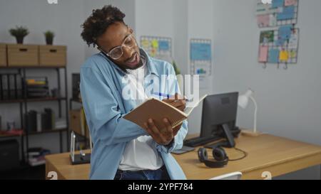Hübscher junger afroamerikanischer Mann, der am Telefon multiagiert, während er Notizen in einer modernen Büroumgebung macht. Stockfoto