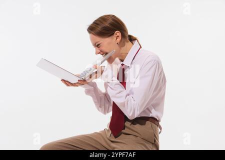 Stilvolle junge Frau in weißem Hemd und roter Krawatte beißt in einem Moment humorvoller Frustration vor weißem Studiohintergrund auf ihren Laptop. Stockfoto