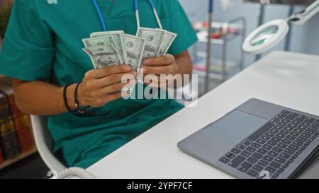 Ein hispanischer Arzt zählt uns Dollarscheine in einer Klinik mit einem Laptop auf dem Schreibtisch in einem gut beleuchteten, organisierten medizinischen Arbeitsplatz. Stockfoto