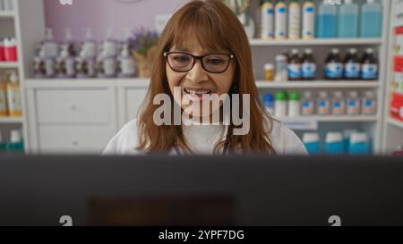 Hispanische Frau mittleren Alters, die in einer Apotheke arbeitet, lächelnd und mit Brille, mit Regalen mit Produkten im Hintergrund. Stockfoto
