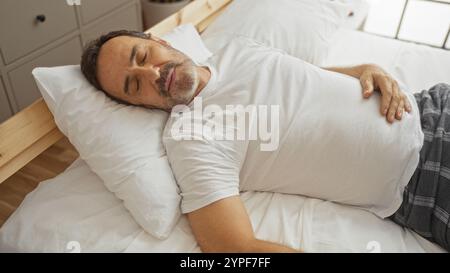 hispanischer Mann mittleren Alters, der friedlich in einem gemütlichen Bett in einem gut beleuchteten Schlafzimmer schläft, bietet Komfort und erholsamen Schlaf in einer häuslichen Umgebung. Stockfoto