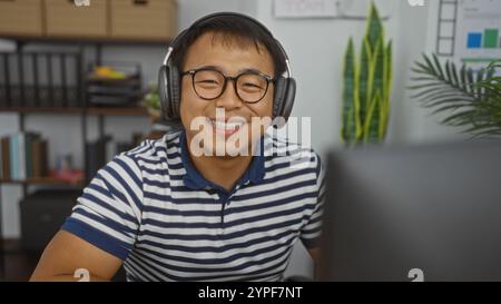Hübscher junger chinesischer Mann, der in einem Büro im Innenbereich Kopfhörer trägt. Stockfoto