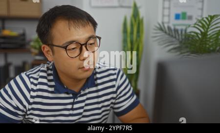 Hübscher junger asiatischer Mann mit Brille und gestreiftem Hemd in einem modernen Büro mit Pflanzen und Diagrammen im Hintergrund. Stockfoto