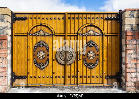 Ein wunderschön gefertigtes Holztor steht vor einem Hintergrund aus Steinmauern. Das Tor verfügt über raffinierte Dekorationen und glänzende Metallgriffe, die von hellem Sonnenlicht beleuchtet werden. Stockfoto