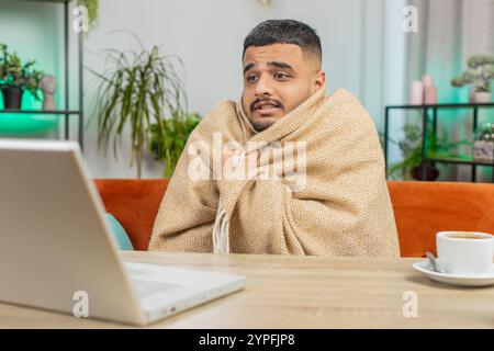 Kranker, kranker indischer Mann, der an einem Laptop arbeitet, der in Karo gewickelt ist, sitzt zitternd vor kaltem trinken heißem Kaffee im unbeheizten Heimbüro, ohne Schulden zu heizen. Ungesunder Araber versuchen, sich aufzuwärmen Stockfoto