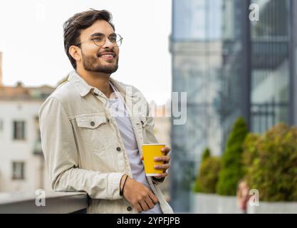 Glücklicher bärtiger indischer Mann, der morgens Kaffee, ein heißes Getränk genießt und draußen lächelt. Entspannen, eine Pause einlegen. Ein arabischer Hindu-Typ in der Stadt, der in der Innenstadt Kaffee trinkt. Stadtleben im Freien Stockfoto