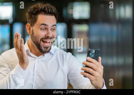 Glücklicher junger Geschäftsmann in weißem Hemd, der am Smartphone arbeitet, schockiert von einem plötzlichen Siegesspiel, das Lotterie-Jackpot-Leistung gewinnt, gute Karrierenachrichten. Aufgeregter Mann im formellen Stehen in der Lobby. Stockfoto