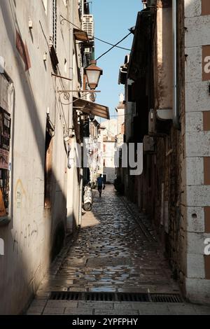 Enge Gasse in der Altstadt von Rovinj, gepflastert mit Kopfsteinpflaster. Touristen laufen entlang der engen Einkaufsstraße. Steingebäude beleuchtet durch Sonnenlicht. Stockfoto